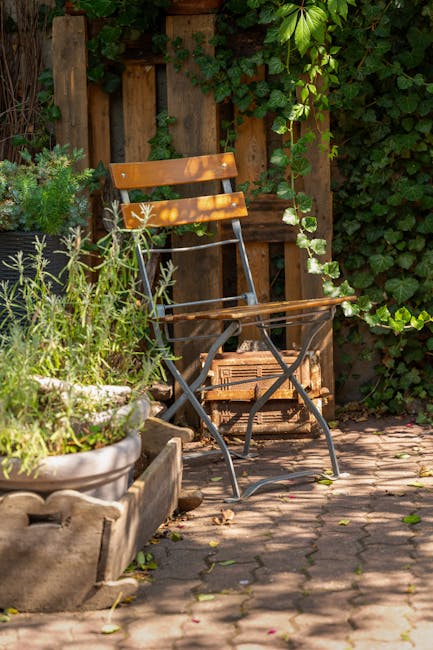 A small urban garden corner featuring a wooden and metal garden chair with a slatted wooden backrest and seat, placed on a cobblestone paved patio. Surrounding the chair, there are lush green plants, including a potted herb or shrub in a large, weathered ceramic container to the left, and various leafy vines and plants climbing a wooden fence behind. The fence is constructed from vertical wooden planks, with some foliage spilling over the top, creating a vibrant, natural backdrop. The light filters softly through the leaves, illuminating the textured soil and the weathered surfaces of the plants and furniture, suggesting a well-maintained outdoor space suitable for relaxing or gardening activities. Gardeners Shoreditch can offer expert garden maintenance and landscaping work to enhance outdoor terraces like this in Shoreditch, London.
