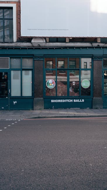 The image depicts the storefront of Shoreditch Balls, a business located on a street with empty paved road in the foreground. The building has a dark exterior with large windows displaying colorful circular signs, and a glass door with signage. The upper part of the building features brickwork and a white panel, suggesting an urban setting. Although the photo primarily captures the storefront, it references the vibrant and diverse environment of Shoreditch near Old Street, an area known for its creative and eclectic character. As part of garden maintenance or landscaping work, external features like the neighboring street and paving are visible, with no plants or gardening tools present. The lighting indicates daytime with soft natural light, emphasizing the urban setting rather than outdoor garden elements.