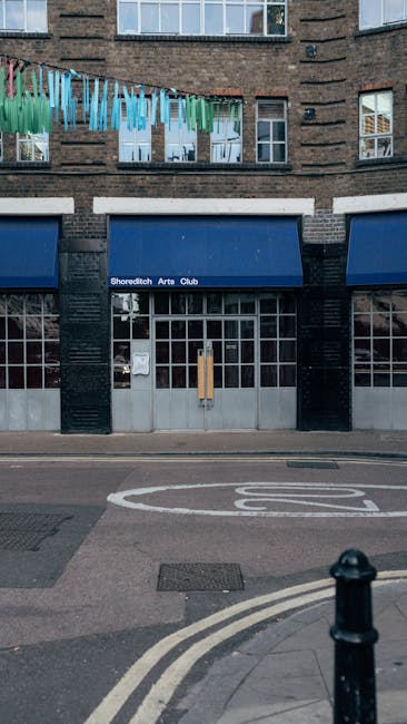 The image depicts the exterior of the Shoreditch Arts Club building, featuring a dark brick facade with three tall, white-framed windows on the upper floors. Above the entrance, a blue sign displays the name 'Shoreditch Arts Club.' On the second floor, a clothesline with colorful garments, including blue, green, and purple fabric strips, is strung across a window, adding a vibrant touch to the scene. The sidewalk in front of the building is paved, with a bicycle lane marked on the road and a black bollard at the street corner. The scene is illuminated by natural light, with reflections visible on the windows. Although the image does not directly feature gardening or outdoor maintenance, the setting is urban with a focus on the building's exterior and street environment, and it is associated with the domain gardenersShoreditch.org.uk, which provides local gardening and landscaping services in this area.